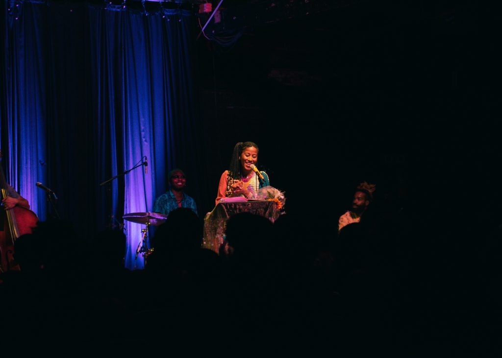 Image: Aja Monet at Constellation, Chicago on September 03, 2023. A  photograph of the poet onstage smiling at an ornate podium. Behind her are several people seated and smiling. The audience's silhouette is visible. Photo credit: Gracie Hammond.