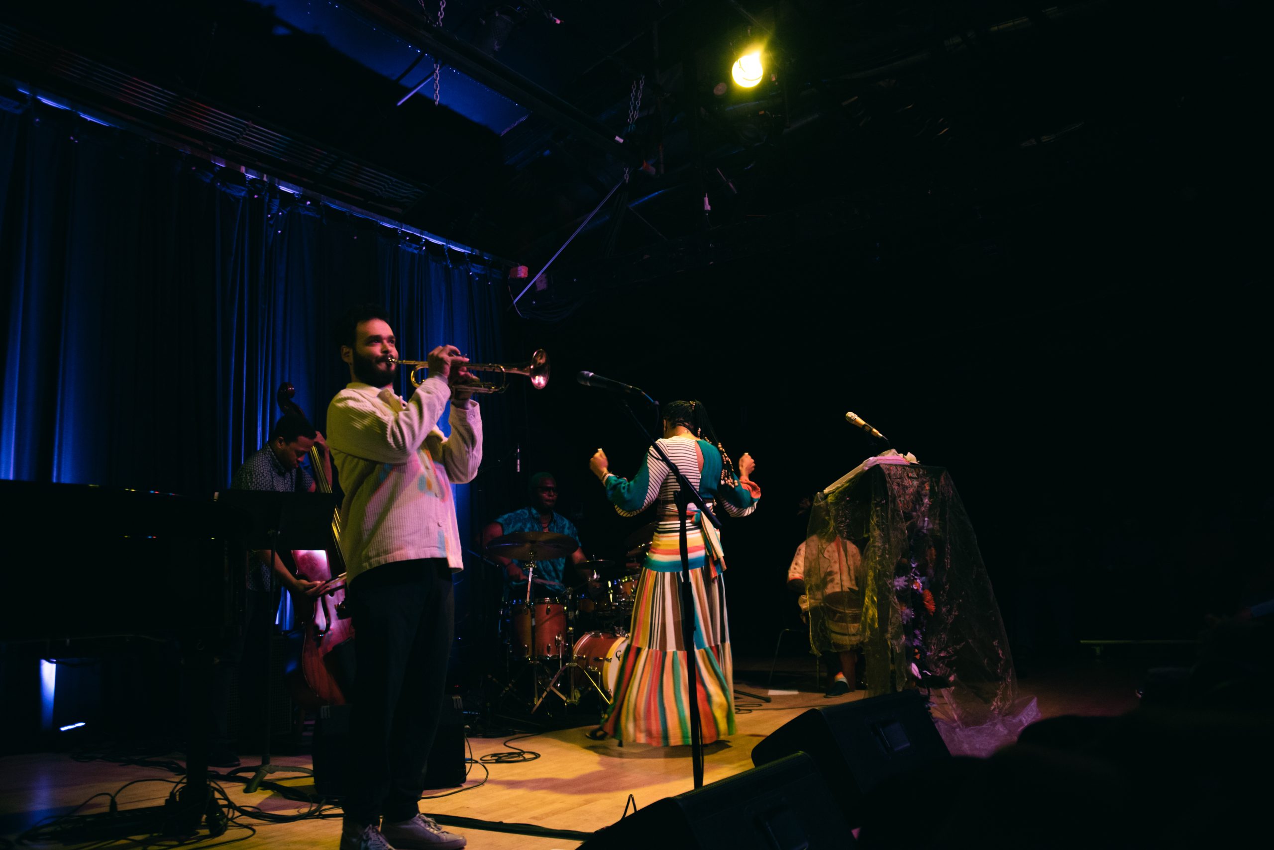 Image: Julian David Reed's trumpet solo at Constellation, Chicago on September 03, 2023. A photograph of the poet onstage with her back turned while Reed, wearing a white sweater, is spotlit and plays the trumpet. Photo credit: Gracie Hammond.