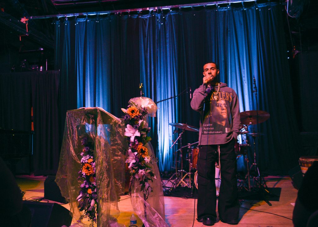 Image: Vic Mensa performing at Constellation, Chicago on September 03, 2023. The rapper onstage wears a graphic sweatshirt, a chain, and black flared cargos next to a floral and fabric installation on the left. Photo Credit: Gracie Hammond.