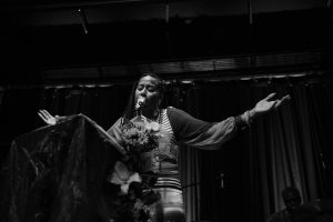 Image: Aja Monet at Constellation, Chicago on September 03, 2023. A black and white photograph of the poet onstage with her arms outstretched and a floral and fabric installation on the left. Photo credit: Gracie Hammond.