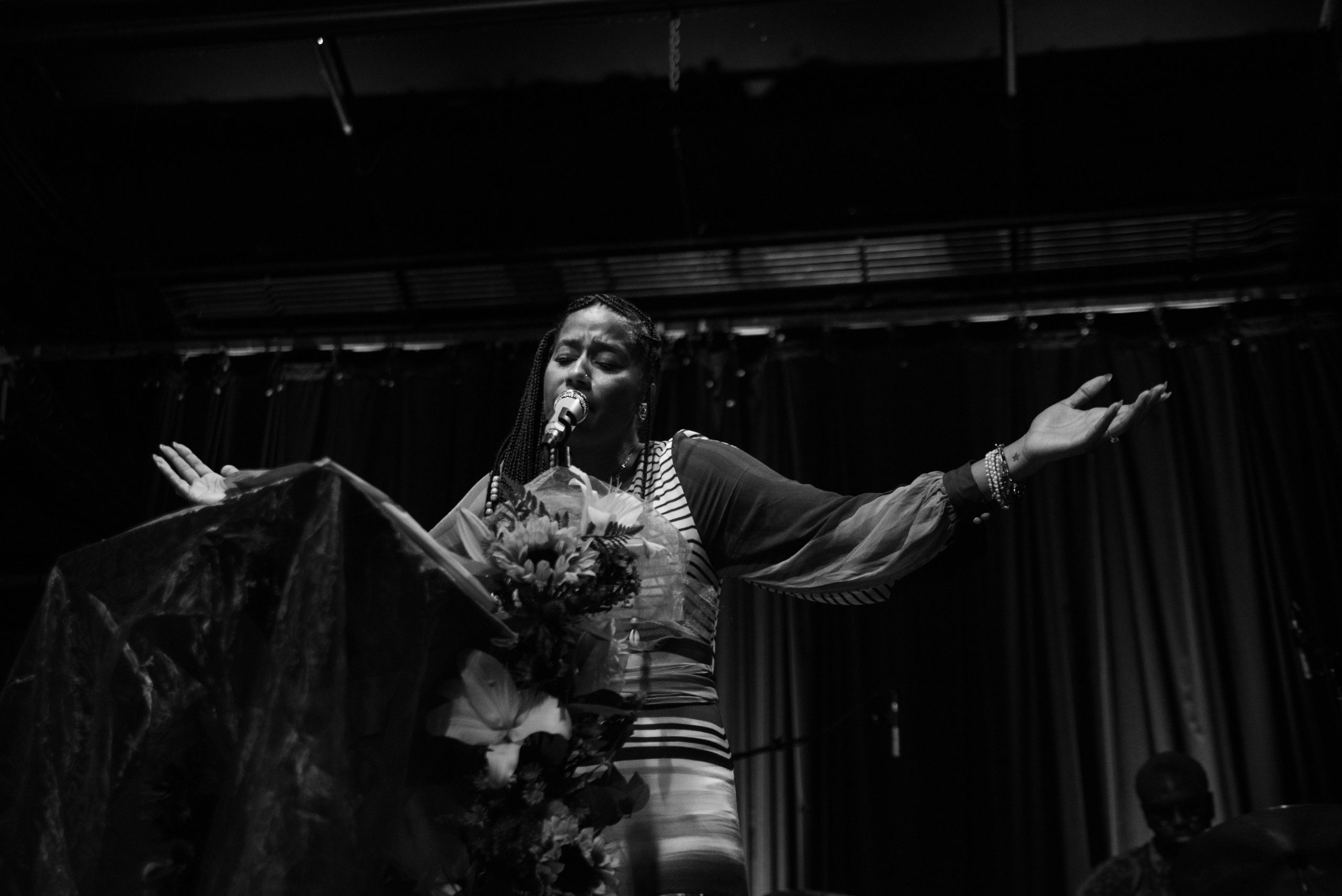 Image: Aja Monet at Constellation, Chicago on September 03, 2023. A black and white photograph of the poet onstage with her arms outstretched and a floral and fabric installation on the left. Photo credit: Gracie Hammond.