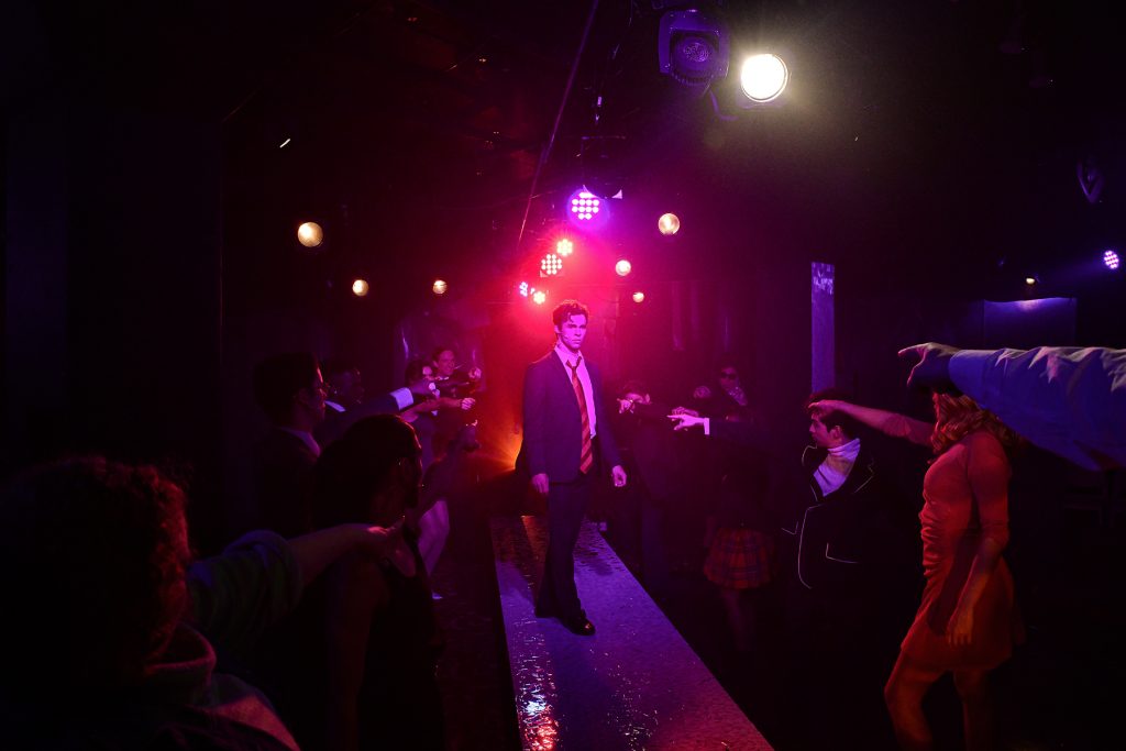 Image: Kyle Patrick stands center of the image on a long platform, wearing a business suit, the blazer open revealing a large tie. The other cast members surround him on either side in lines, pointing at him. Red and purple lights flood the stage. Image by Evan Hanover.