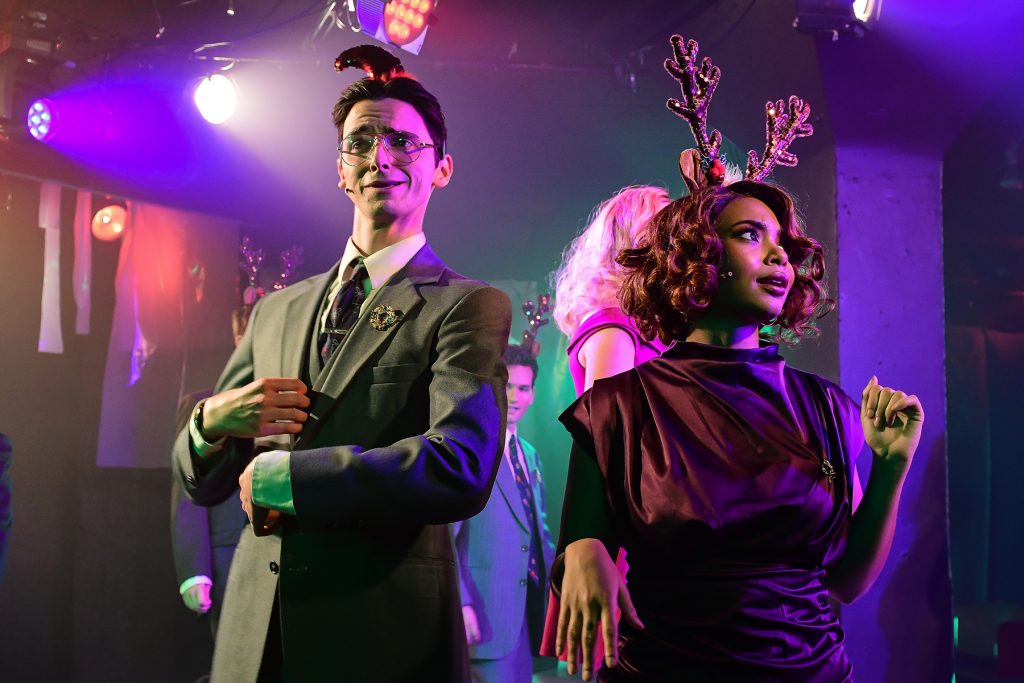 Image: "American Psycho" actors Quinn Kelch and Danielle Simmons stand next to each other, photo capturing them from the waist up. Kelch wears a green business suit with a wreath on their lapel, some wire-rimmed glasses, and a Christmas elf hat. Simmons, looking off-camera to the right, wears a black formal dress and kitschy reindeer antlers. Behind them are other cast members wearing the same kind of reindeer antlers. Photo by Evan Hanover.