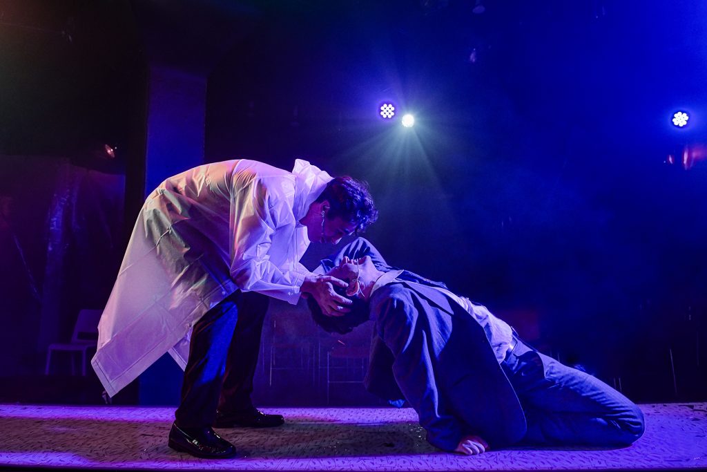 Image: Kyle Patrick bends down to hold John Drea's head and stare into his face; Drea is on his knees leaning his body back. Patrick wears a white plastic coat. Drea wears a suit. Behind them, a blue stage light shines over them. Photo by Evan Hanover.