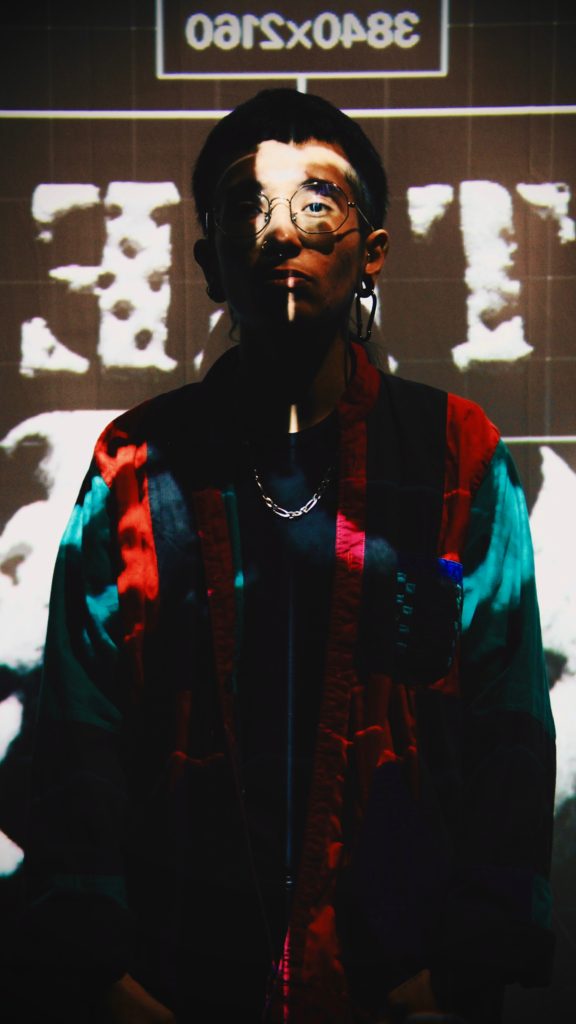 Image: Eme looking serious behind the projection screen as part of the production Tambo & Bones: The Escape at the Den Theatre. Foto x Luz Magdaleno Flores, 2023.