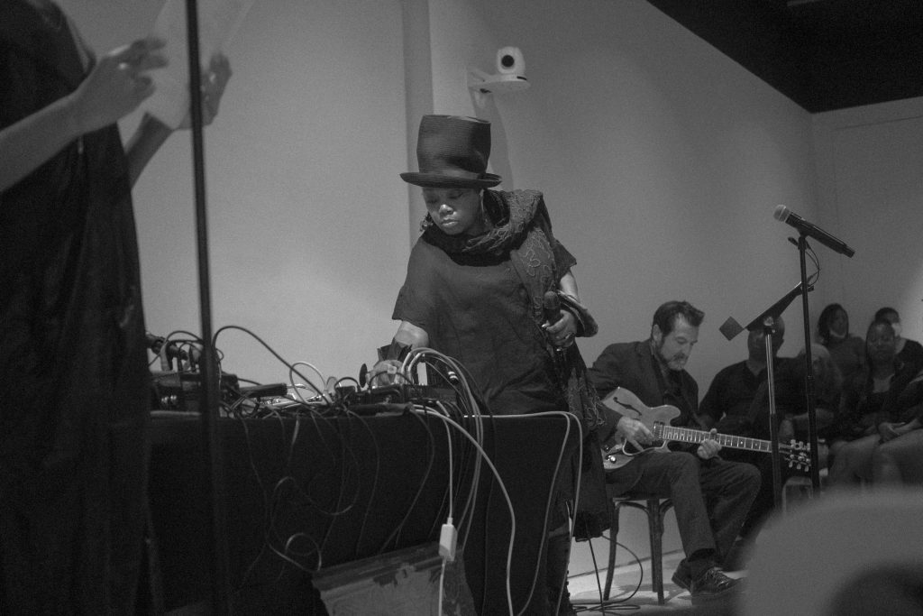 Image: Image: A black and white photograph Dee Alexander & John McLean perform on the Poetry Foundation’s stage during Herman’s Lounge. Alexander is wearing a dress and scarf with a top hat and tinkering with a sequencer. Rucker is reading poetry at mic. A guitarist and audience member's sit in chairs to their left. Photo by Oriana Koren.