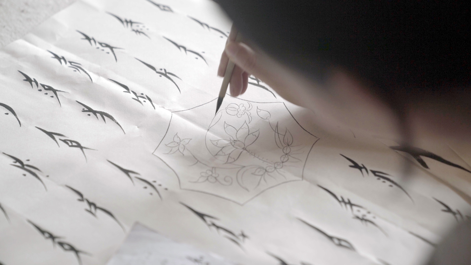 Image: Hu Xin's handwriting in Nüshu. Still from "Hidden Letters," 2022, directed by Violet Du Feng and Qing Zhao. A close-up in the film that shows the writing system "Nüshu." A right hand is seen holding a calligraphy brush. On the rice paper, vertical lines of slender characters, illegible to most, elegantly frame a octagon motif featuring a line drawing of a lotus. Photo credit: Jia Li.