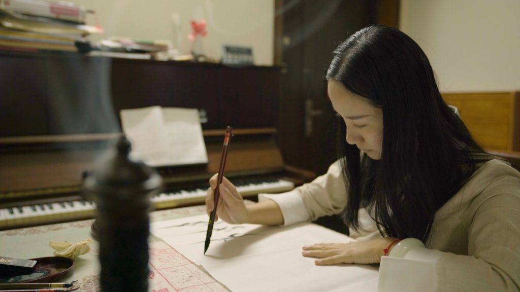 Image: Simu writes Nüshu. Still from "Hidden Letters," 2022, directed by Violet Du Feng and Qing Zhao. A young Chinese woman is seen, sitting in a private room, concentrating on her practice of Nüshu using a traditional calligraphy brush and writing on rice paper. Her long black loose hair frames her face and covers part of the traditional Chinese attire she's dressed in. She wears a jade bracelet, as well as a red string bracelet, on her left wrist. There seems to be an incense burner in the foreground and a piano in the background; both are out of focus. Photo credit: Feng Tiebing.