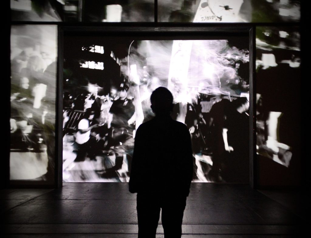 Image: Eme's black shadow standing in front of a projection as part of the production Tambo & Bones: The Escape at the Den Theatre. Foto by Luz Magdaleno Flores, 2023.