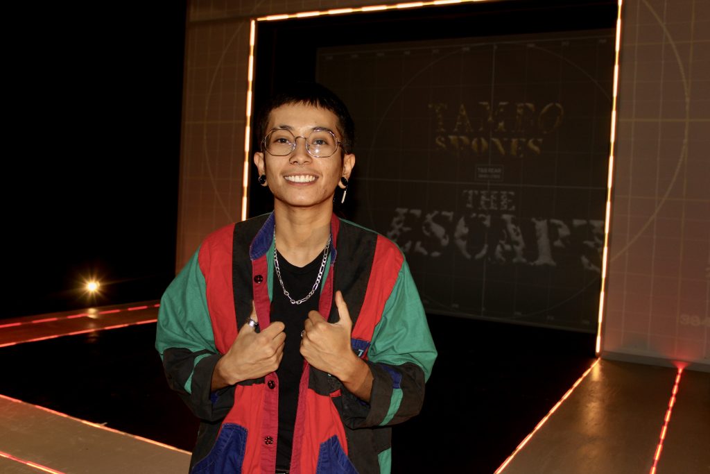 Image: Eme standing in front of the stage wearing a red, black, green, and blue jacket and silver chain with a projection behind them that reads, "Tambo & Bones" at the Den Theatre. Foto by Luz Magdaleno Flores, 2023.