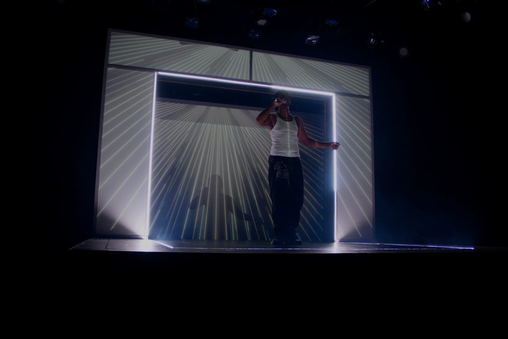 Image: Tambo played by William Anthony Sebastian Rose II in front of a projection of shining light beams by Eme as part of the production Tambo & Bones: The Escape at the Den Theatre. Foto by Luz Magdaleno Flores, 2023.