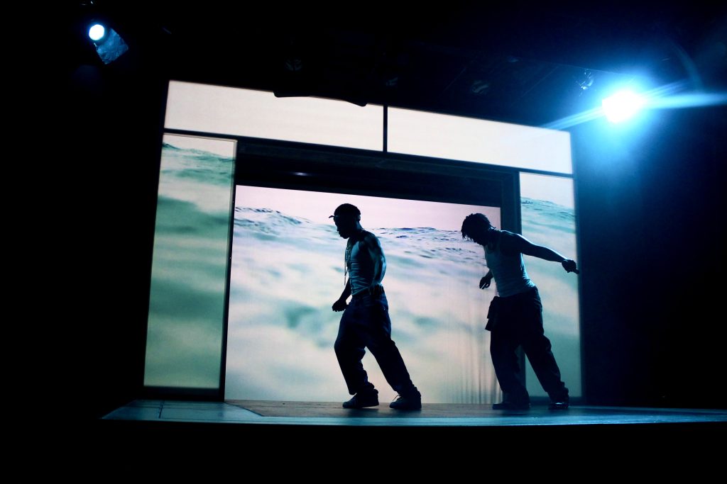 Image: Tambo played by William Anthony Sebastian Rose II, and Bones played by Patrick Newson Jr. in front of a projection of a body of water designed by Eme as part of the production Tambo & Bones: The Escape at the Den Theatre. Foto by Luz Magdaleno Flores, 2023.