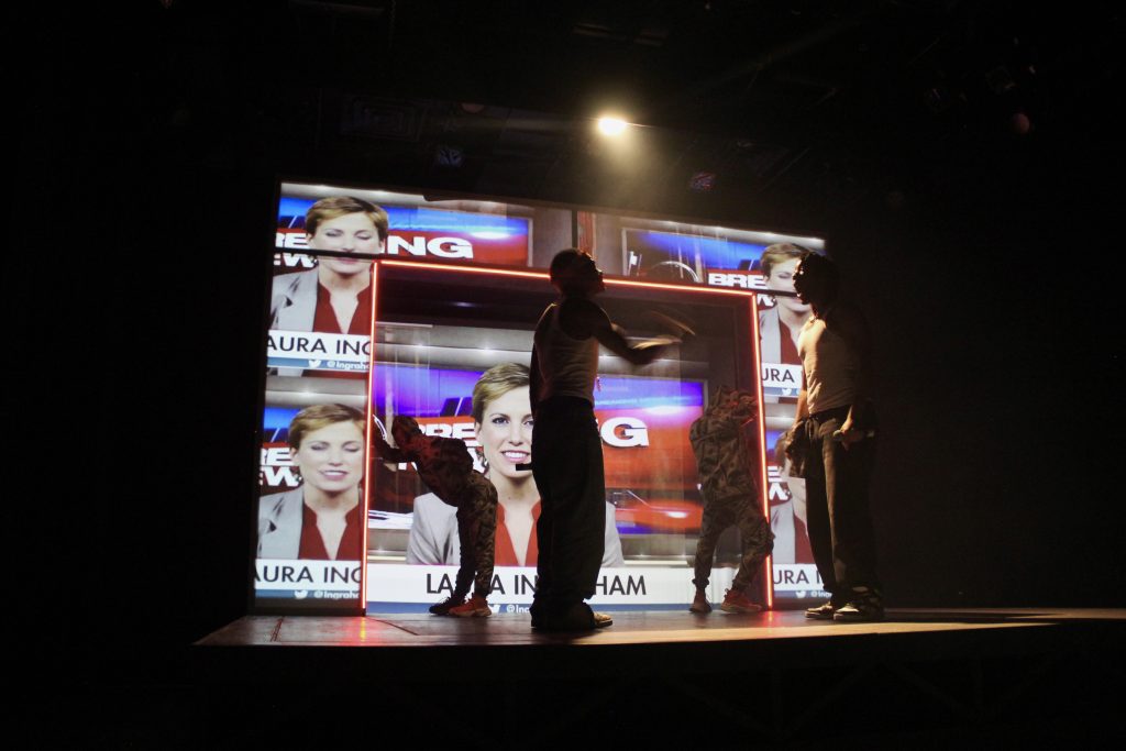 Image: Tambo played by William Anthony Sebastian Rose II, and Bones played by Patrick Newson Jr., and background dancers in front of a projection of a newscaster made by Eme as part of the production Tambo & Bones: The Escape at the Den Theatre. Foto by Luz Magdaleno Flores, 2023.