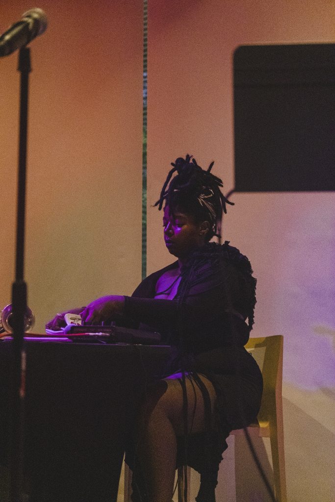 Image: KeiyaA, dressed in a black ensemble and a braided updo, conducts a tarot reading while seated on the stage during Herman's Lounge. The background of the stage is white with a still image projected. Photo by Oriana Koren