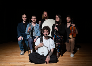 Image: Professional photographic portrait of all of O Death's cast members each holding their instruments on the stage of Facility Theater. All members are standing expect for Quintana who is seated cross-legged in front holding a banjo. Image courtesy of Santiago Quintana.
