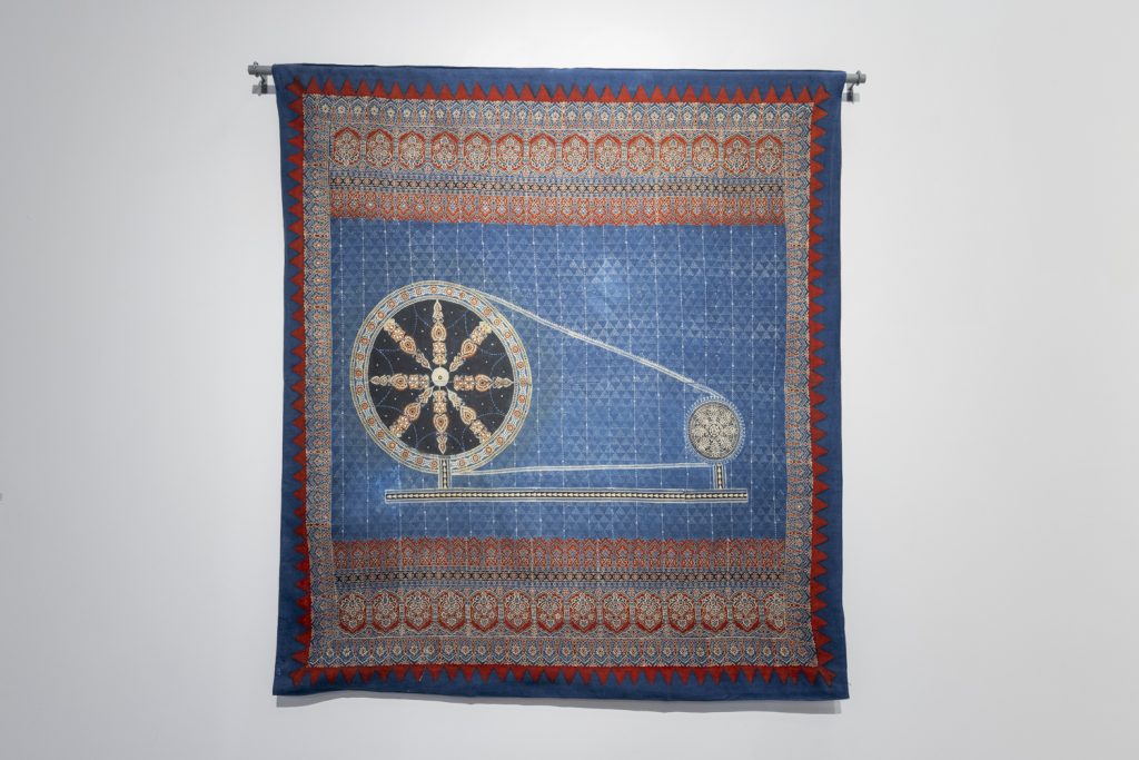 Image: Chakra, 2023. Indigo-dyed wall tapestry. A spindle-like image appears in the center with decorative red, white, and blue details around the edges. Courtesy of the South Asia Institute.