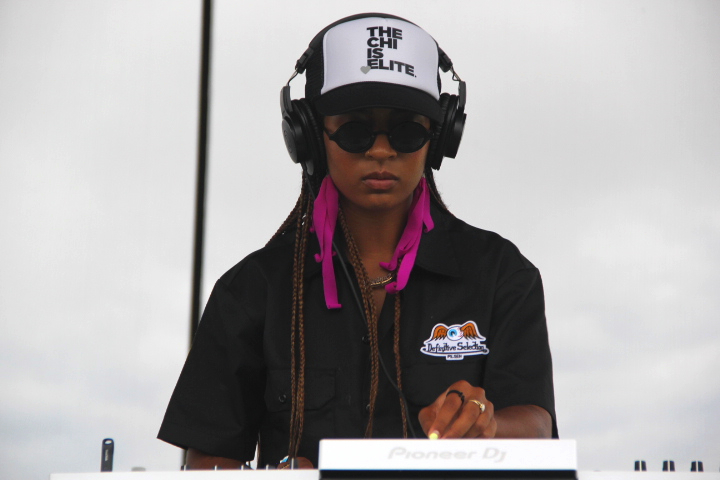 Image: A DJ wearing a hat that says The Chi Is Elite and black headphones over their braided hair and a black button up that has a logo for Defnitive Selection performing their set at My House Music Festival. Foto x Nancy David Sánchez Tamayo, 2023.