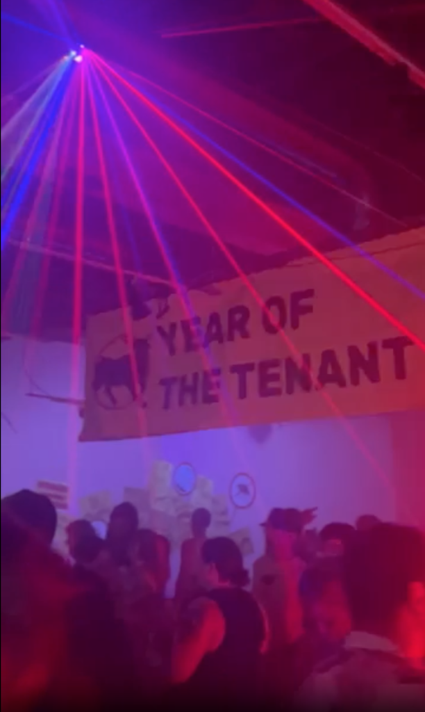 Image: A banner reading “Year of the Tenant” hangs above crowd of partygoers. Red and blue beams of light shine onto the crowd from the ceiling. Photograph by Christina Ostmeyer.