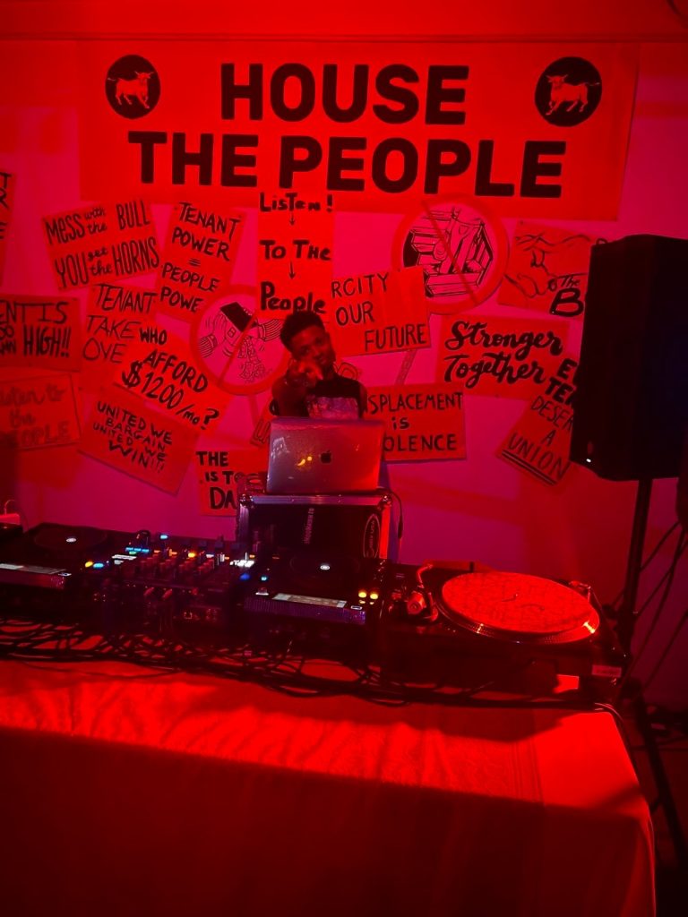 Image: A DJ named Bazzie points at the camera from behind a DJ booth. A large banner reading “House the People” is on the wall above many smaller posters with phrases such as, “Our City, Our Future,” “United We Bargain, United We Win,” and “Displacement is Violence.” Photograph by Christina Ostmeyer.