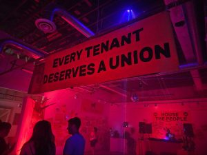 Image: Interior shot of the House the People party. A banner reading “Every Tenant Deserves a Union” is hanging across the upper portion of the photograph. People in the foreground mingle and look at art on the walls. A DJ booth is in the background. The entire scene is lit by red and blue lights. Photograph by Cult Mayor.