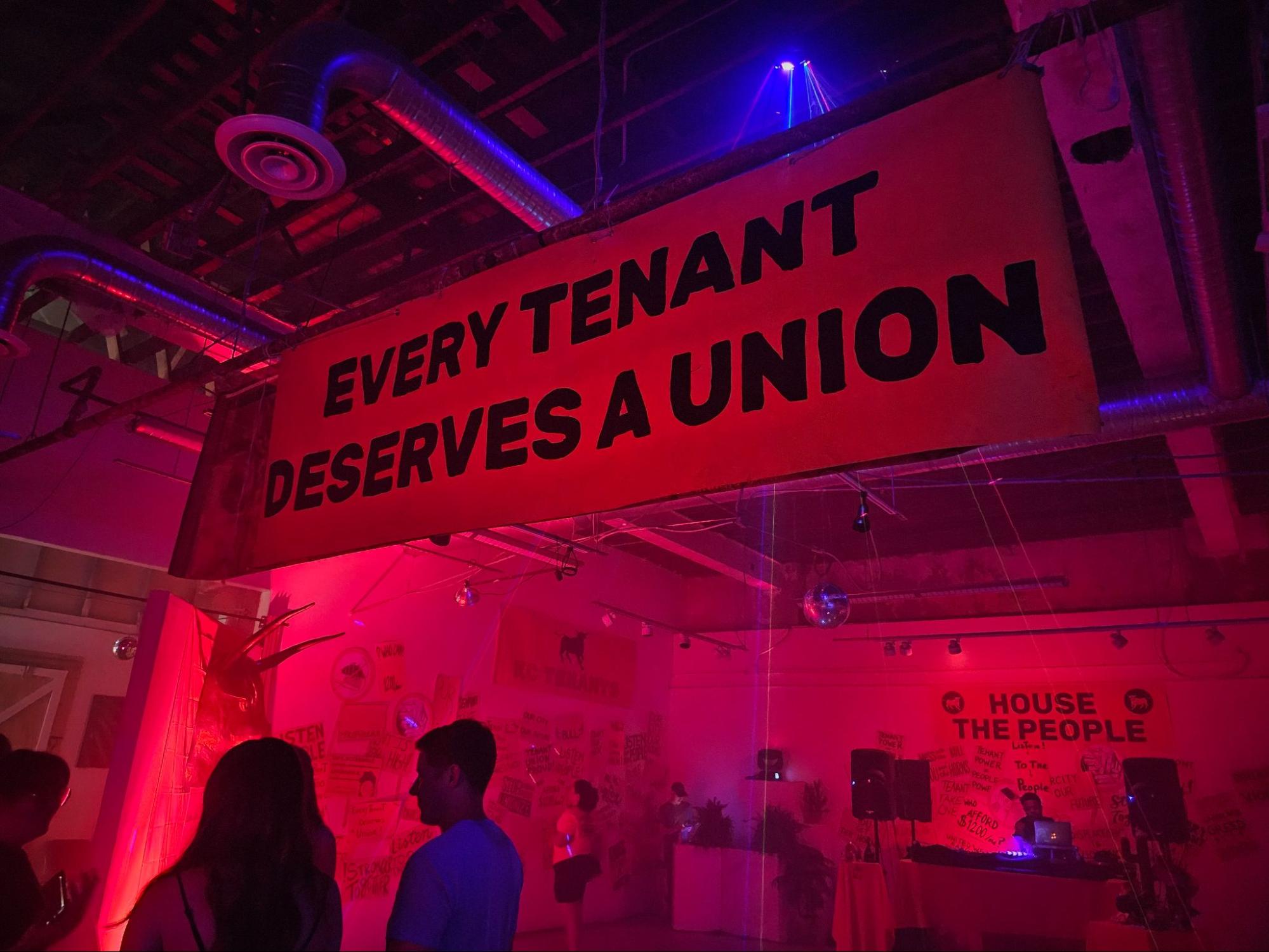 Image: Interior shot of the House the People party. A banner reading “Every Tenant Deserves a Union” is hanging across the upper portion of the photograph. People in the foreground mingle and look at art on the walls. A DJ booth is in the background. The entire scene is lit by red and blue lights. Photograph by Cult Mayor.