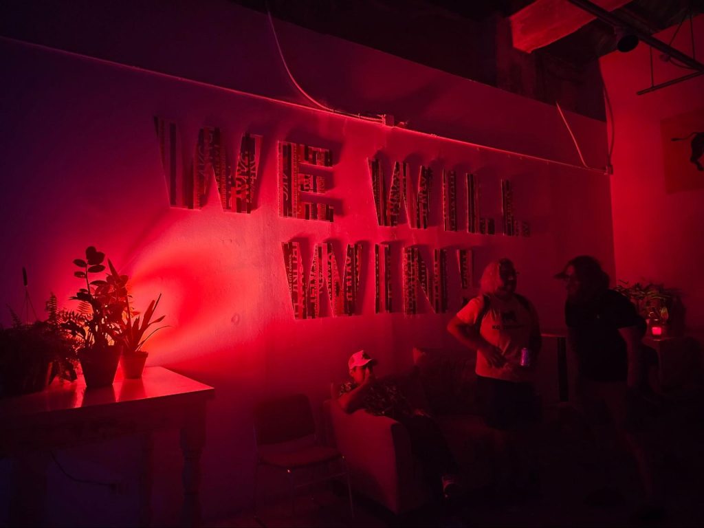 Image: A wall installation of the phrase “We Will Win!” The installation is lit from the left side by a red light. Partygoers chat and mingle in front of the installation. Photograph by Cult Mayor.