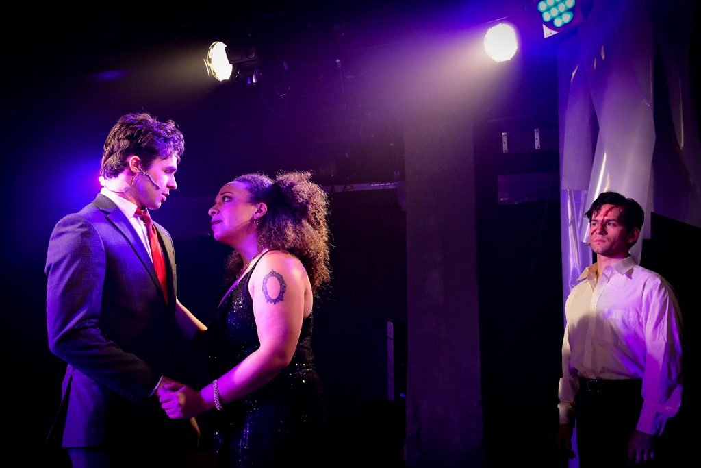 Image: On the left side of the image, Kyle Patrick and Sonia Goldberg stand in-profile looking at each other. Patrick wears a business suit and tie, Goldberg wears a sequined night dress. To the right, John Drea looks at the other two, wearing a white button up; on his face is a long red cut. Photo by Evan Hanover.