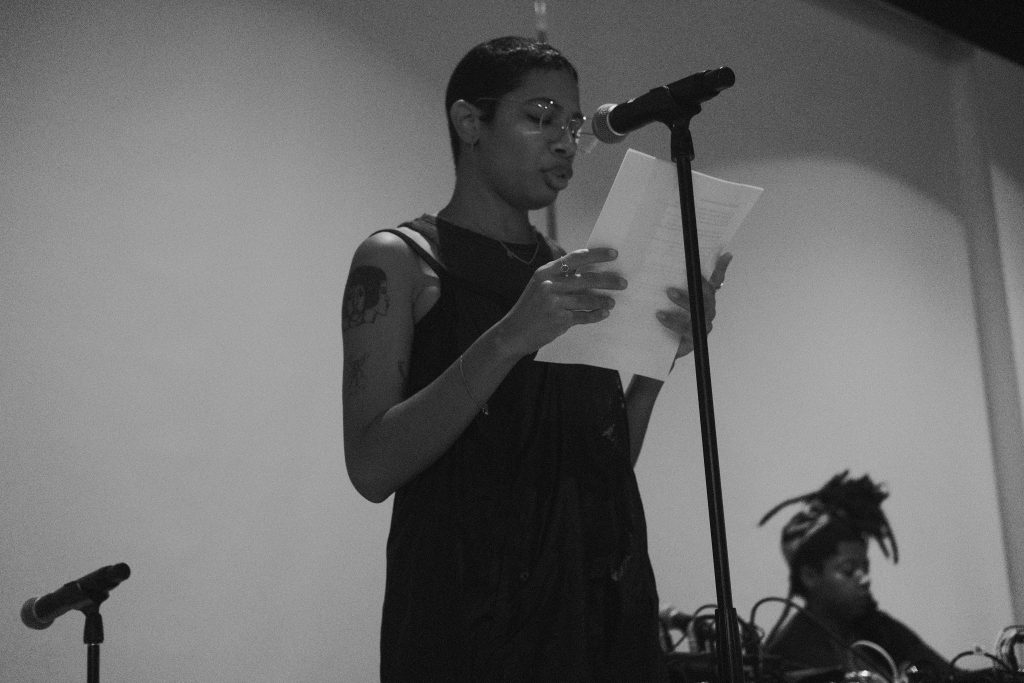 Image: Gabrielle Octavia Rucker reads their poetry on the stage of the poetry Foundation. Rucker is a wearing black dress, jewlery on their wrists and fingers, and circular glasses. Artist KeiyA sits playing electronic music off to Rucker's left side. Photo by Oriana Koren.