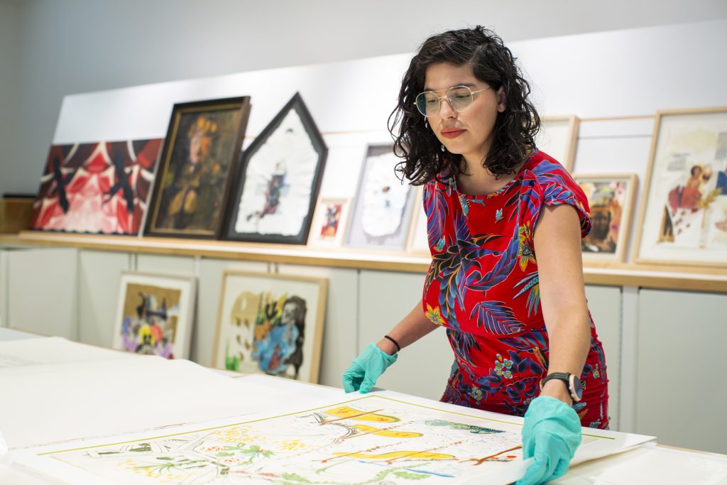 Image: Natasha Mijares conducting research in the collection of Latinx art at DePaul Art Museum. She is holding the edges of a large piece of art sitting on a table. She is wearing a red dress with a floral pattern and teal gloves. Photograph by Kristie Kahns.