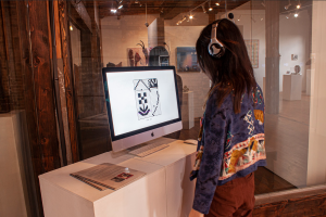 Image: [Fig. 1] On-site view of “Woven Symphony” at Woman Made Gallery; a person wearing headphones stands at in front of a white plinth looking at a computer monitor with a white and black grid pattern in the center of it. Photo by Qianwen Yu.