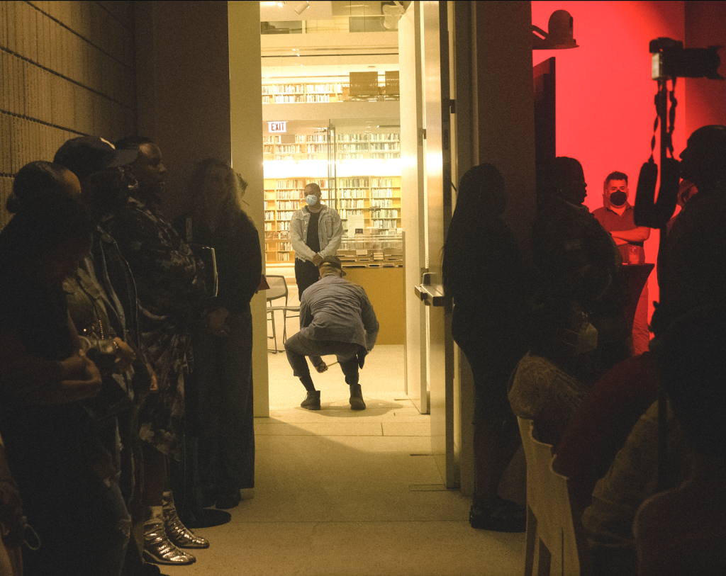  Right Image: An image of the Poetry Foundation entryway to the performance space during Herman's Lounge. Ben Lamar Gay is faced away from the camera. and is squatting in the middle of the doorway as part of a movement piece. Onlookers are standing against the wall behind the seated audience members. A soft red light emanates from the performance space whilst yellow light comes in through the entryway, which reveals a library stacked full of books. Photo by Oriana Koren.