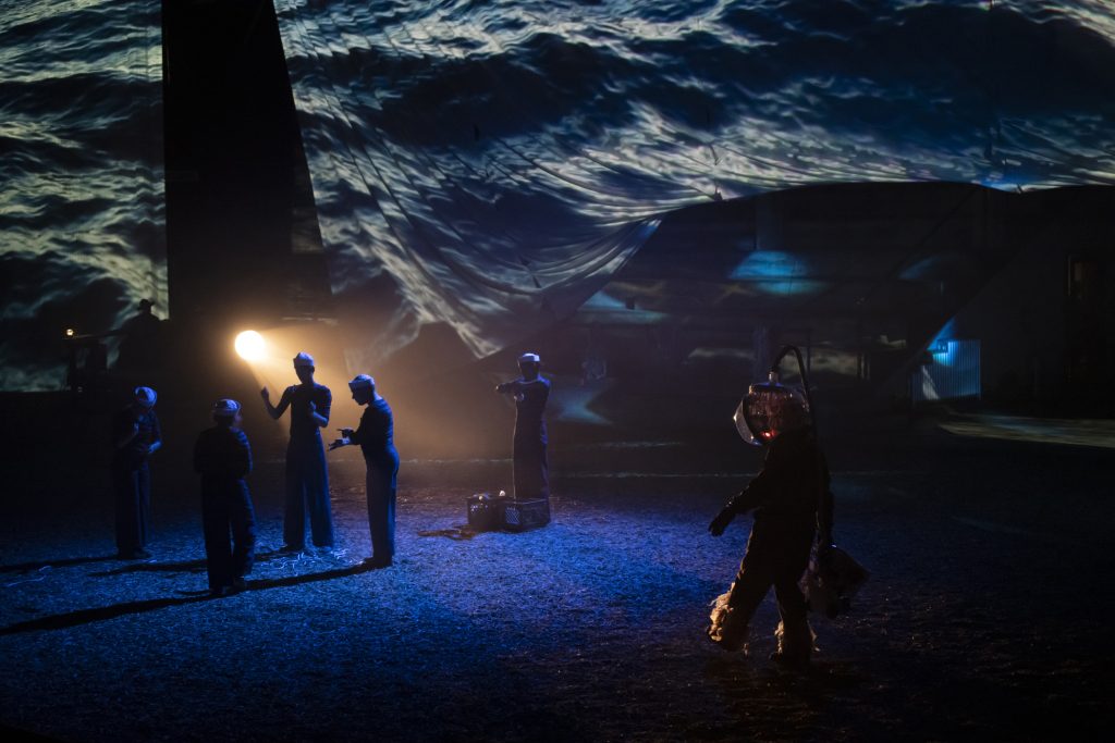 Image: Scene from CETACEAN, staged at the University of Illinois Stock Pavilion, September 27-October 2, 2023. Very dramatic lighting sets the stage for four actors dressed as sailors, and one person wearing an aquanaut costume with a large, glass helmet. There is one warm-colored spotlight illuminating the pathway of the aquanaut. Behind the actors is a billowing screen upon which it appears images of the ocean surface are being projected. Photo by Nathan Keay, courtesy of Deke Weaver.