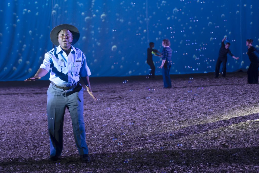 Image: Scene from CETACEAN, staged at the University of Illinois Stock Pavilion, September 27-October 2, 2023. Actor Latrelle Bright, a Black woman, is dressed as a “ranger,” with khakis and a light colored collared shirt and a brimmed park ranger hat. She is mid-sentence. Behind her there are four dancers, three of whom appear to be wearing black, and one wearing blue. There are actual bubbles all around them, and oceanic bubbles projected on the screen behind them. Photo by Nathan Keay, courtesy of Deke Weaver.