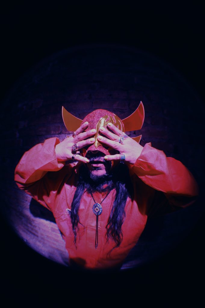 Image: Chébaka in a red Luchado mask. He is wearing a read shirt and a bolo tie. His hands cover his eyes with his fingers adorned with many rings. Photo by Luz Magdaleno Flores.