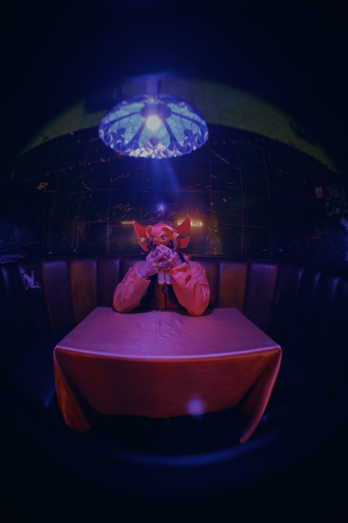 Image: Chébaka sits at a table with a red cloth. His hands hold up a little doll over his face. The doll matches his outfit, which includes a red shirt and a red Lucha mask with gold flames. Photo by Luz Magdaleno Flores.