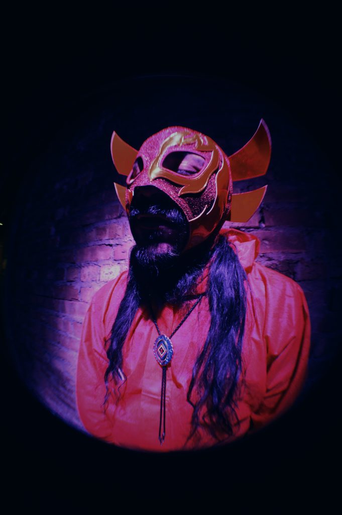 Image: Chébaka stands in front of a brick wall while wearing a red Luchador mask that has gold flames on it. He is also wearing a read shirt and a bolo tie and his eyes are closed. Photo by Luz Magdaleno Flores.