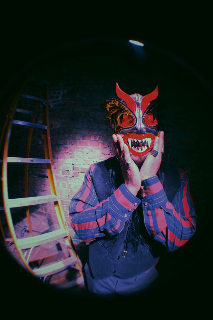 Image: Chébaka stands in front of a brick wall next to a ladder wearing a striped shirt, vest, bolo tie, and several rings. He wears a Luchador mask that is black and red. His hands are on either side of his face. Photo by Luz Magdaleno Flores.