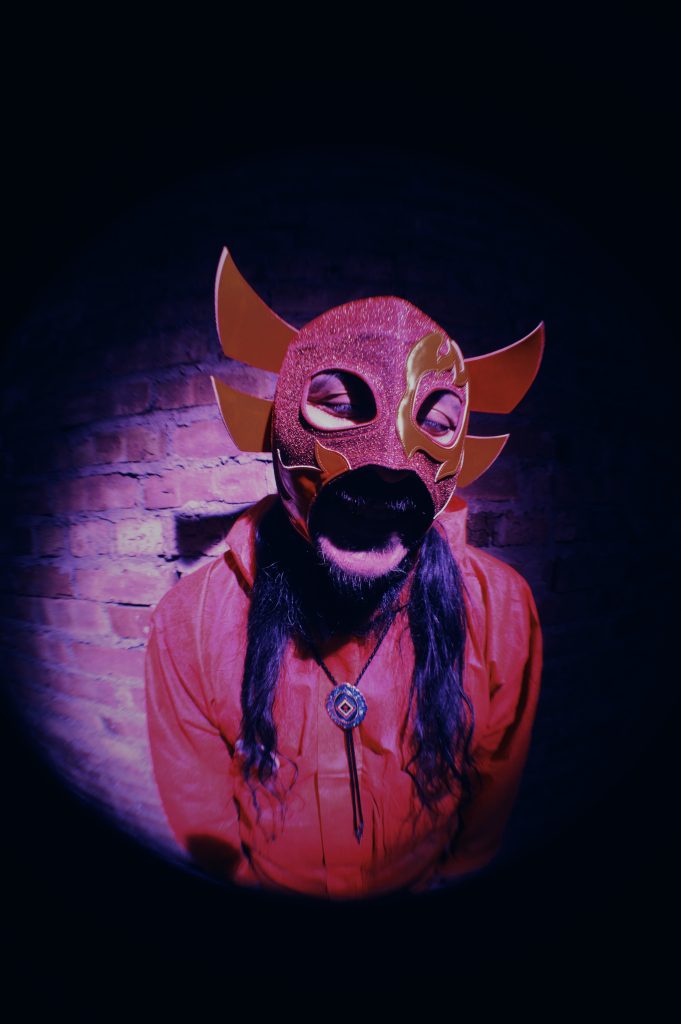 Image:  Chébaka stands in front of a brick wall while wearing a red Luchador mask that has gold flames on it. He is also wearing a read shirt and a bolo tie and his eyes are closed. Photo by Luz Magdaleno Flores.