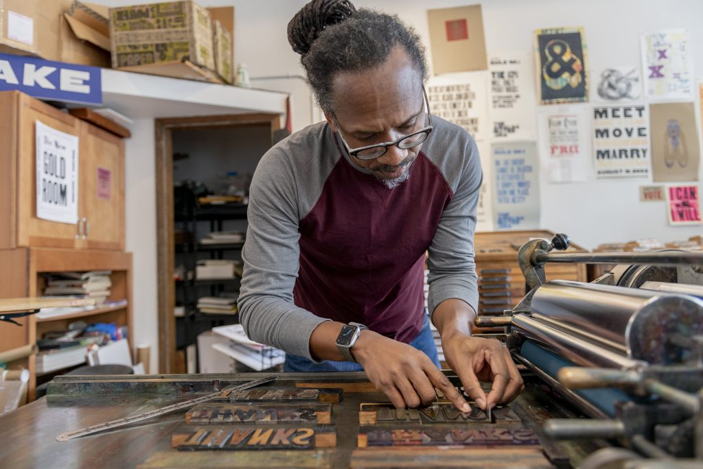 Image: Ben Blount is in his studio using his letterpress. His hands are lining up wood type, which sit on his printing press. He is wearing a gray and maroon shirt and glasses. Behind him are various posters on the wall. Photo by Ryan Edmund Thiel.