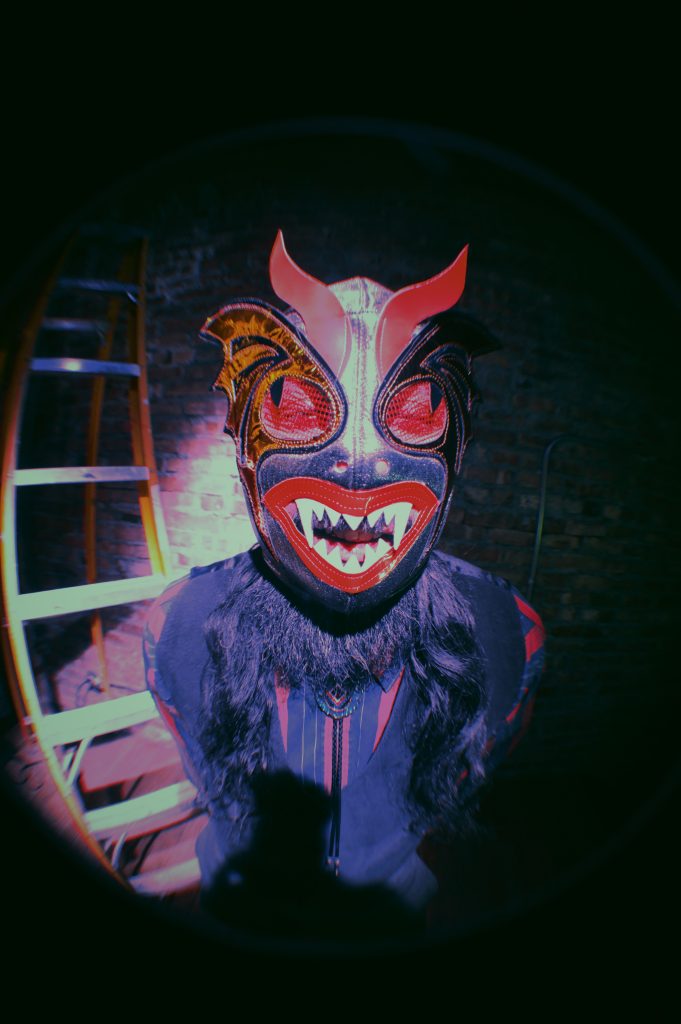 Image (right): Chébaka stands in front of a brick wall next to a ladder. He wears a striped shirt, vest, bolo tie, several rings and a Luchador mask that is black and red. His hands are behind his back as he leans forward towards the camera.