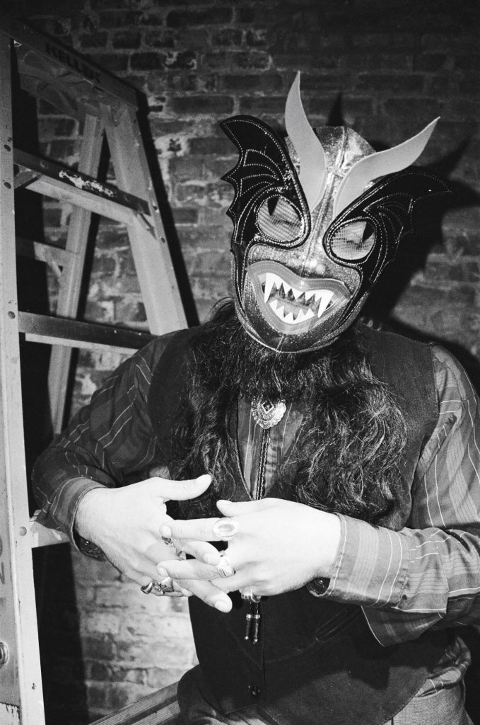 Image: A black and white photo of Chébaka standing in front of a brick wall next to a ladder wearing a striped shirt, vest, bolo tie, and several rings. He wears a Luchador mask and looking towards the camera. His hands are together as if in mid-conversation. Photo by Luz Magdaleno Flores.