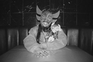 Image: A black and white photo of Chébaka sitting at a table. he wears a long-sleeve shirt and a Lucha mask. He is holding a little doll that wears the same outfit. Photo by Luz Magdaleno Flores.