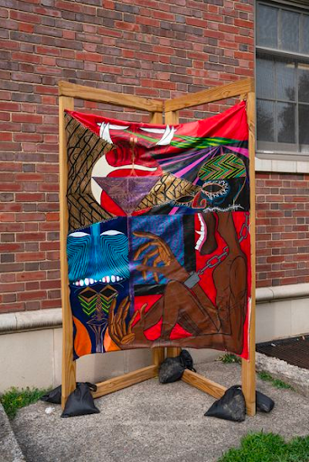 Jamaa Gee presents a red, brown, blue, purple, colorful patterns on a unique tapestry that hangs on a wooden structure being held down by black sand bags. 