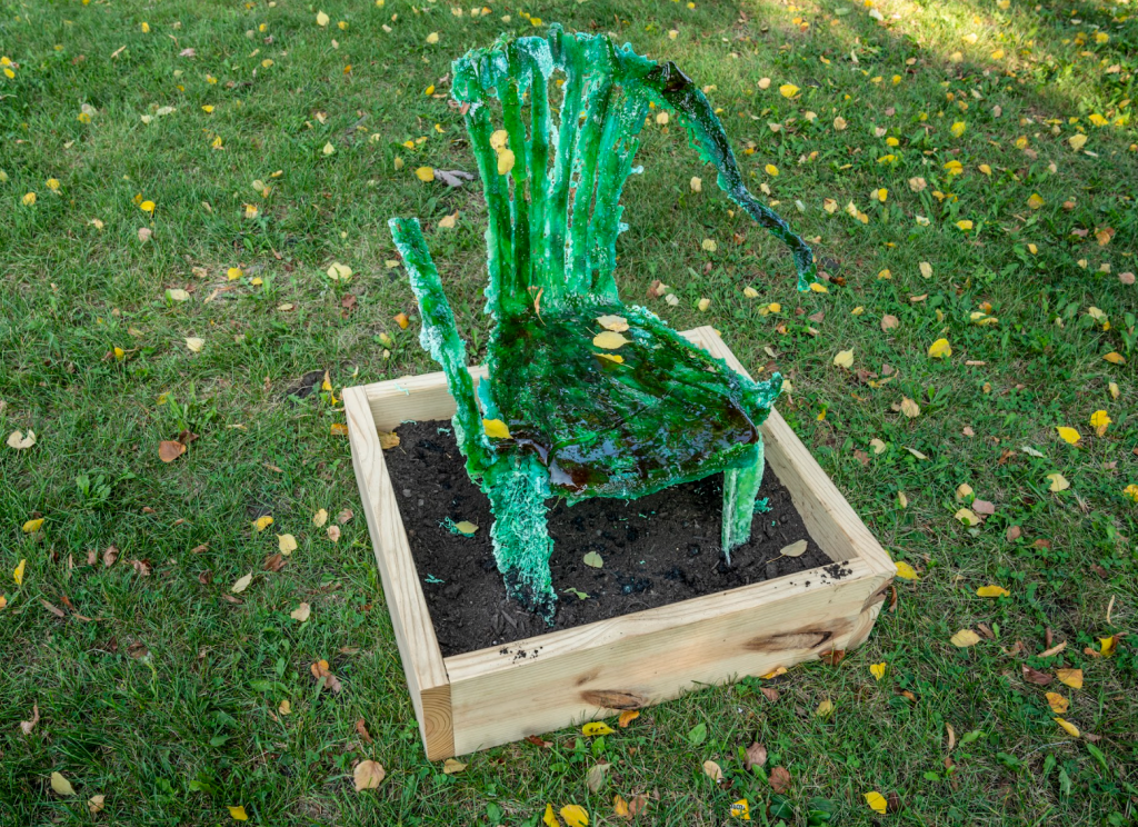A green sculpture of a melting chair by Tavia David sits in a dirt box outside in a green grass section of the park. 
