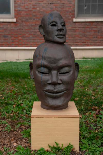 Sculpture of a smaller dark brown human head sits on top of another bigger human head of the same color. Both faces have a dark blue paint drip on them. Presented by Kenya Cree.