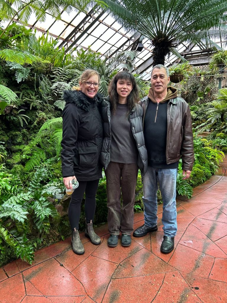  Image: Dorothy and her parents smile and pose together at Lincoln Conservatory Fern Garden during the tenure of her installation, each wearing dark winter coats, boots and jeans. Behind them is lush greenery. They stand on a red tiled pathway. From left to right: Kirsten Carlos, Dorothy Carlos and Felix Carlos. Image by Dorothy Carlos.