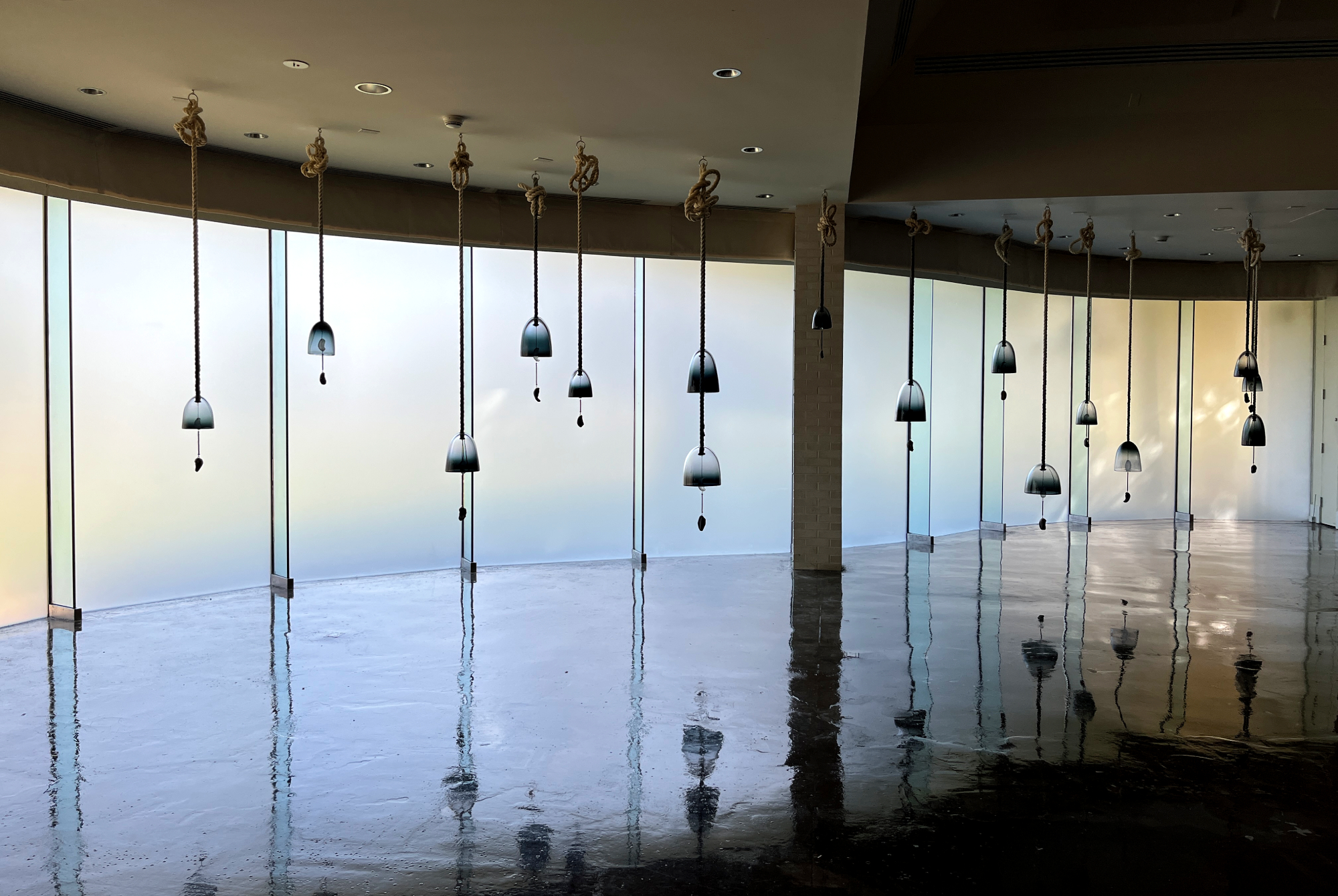 Image: Jenny Kendler and Andrew Bearnot, Whale Bells, 2019-ongoing, hand-blown ombré glass, miocene-era fossilized whale ear bones, cotton rope, and felt. Installation in a museum space with a frosted glass paneled wall as a backdrop. From the ceiling hang approximately fourteen ropes at the bottom of which are glass bell shapes. From the bells hang fossilized whale ears. The light is dramatic, and the bells are reflected in the shiny floor. Photo by Jessica Hammie.