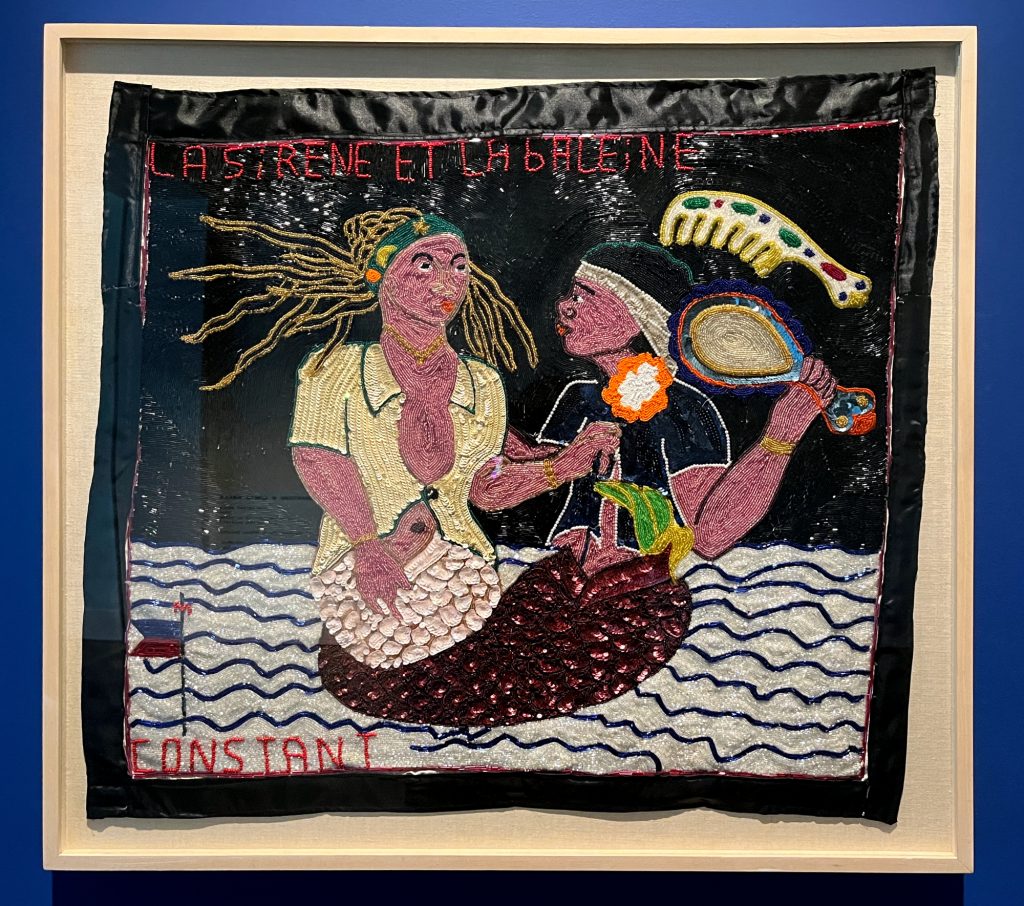 Image: Myrlande Constant, La Sirene et la Baleine, c. 2000, sequined tapestry (drapo Vodou). The tapestry is framed in a white shadowbox. The background of the tapestry is black, with two light reddish-brown half-human, half-fish/whale figures entangled in the middle. The figure on the left has long yellow hair and is wearing a light yellow collared shirt with one button in the middle. The figure on the right is holding a mirror and has a large, bejeweled comb in their hair. They are pictured on a white and blue striped “ocean.” Constant is in red sequins on the bottom left. Photo by Jessica Hammie.