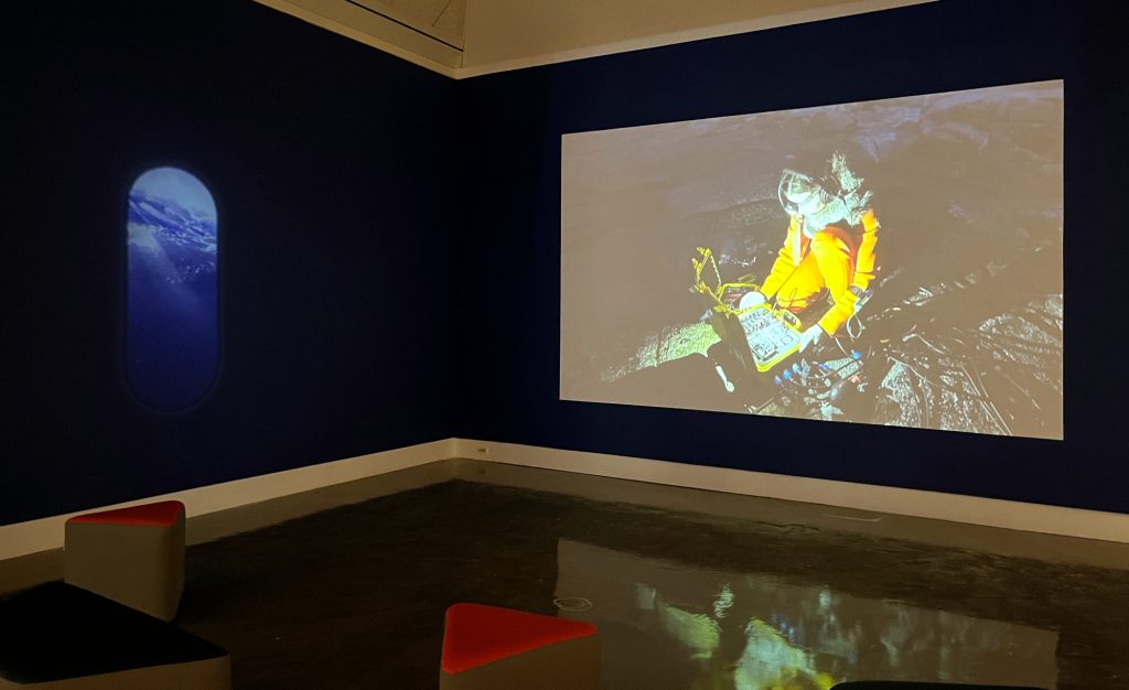 Image: Installation view of Ursula Biemann, Acoustic Ocean, 2018, video installation. On two adjacent walls painted a dark navy blue are two projections. On the left, a projection in the shape of a rounded-top window, like on a plane or submarine. The image projected appears to be the ocean. On the right, a rectangular projection of a scientist wearing a yellow-orange jumpsuit investigates scientific equipment on a rocky shoreline. It is dark, so the only light is from a headlamp that is illuminating the equipment. Photo by Jessica Hammie.