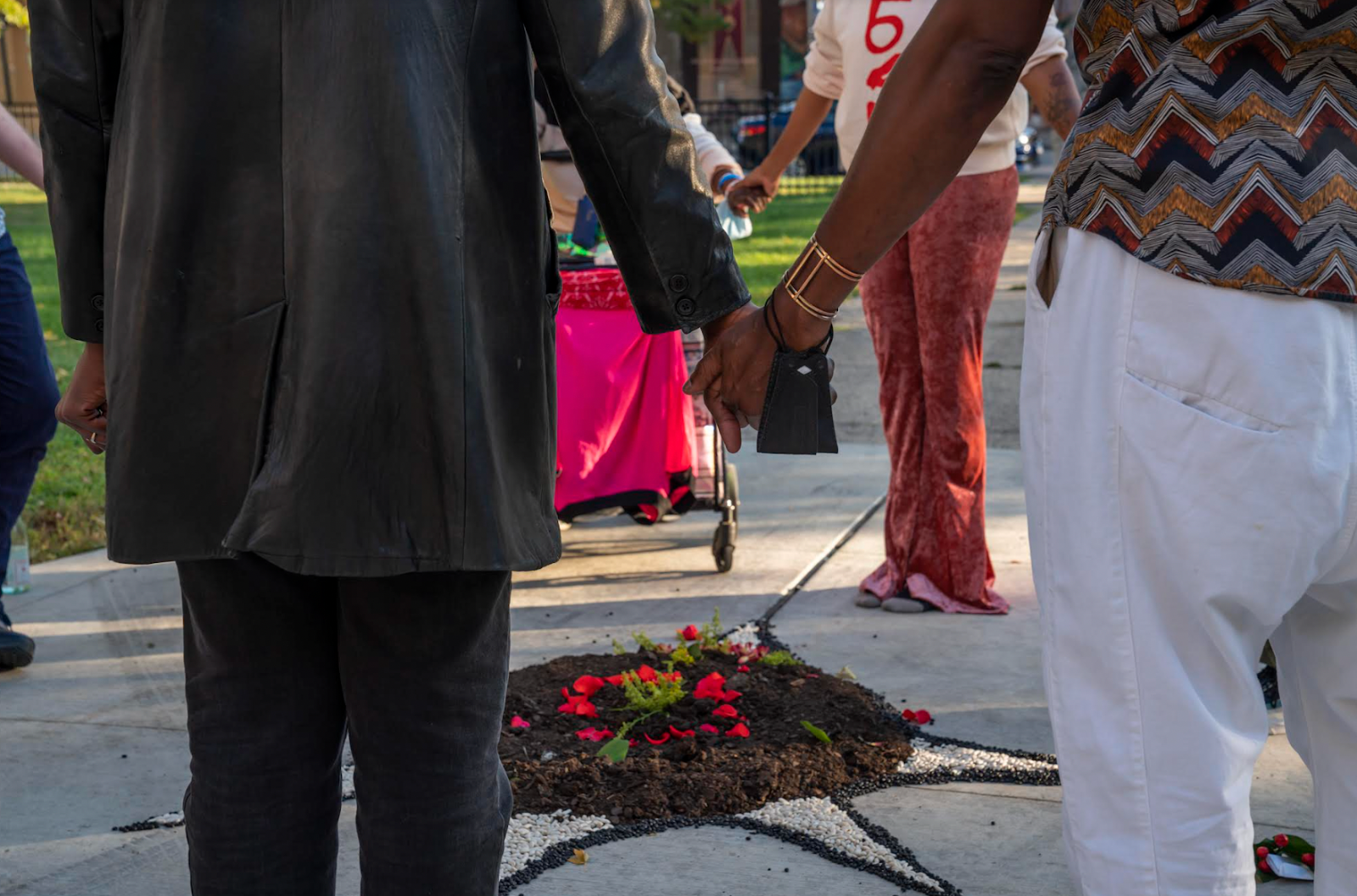 Image: A circle of people holding hands around a floral and dirt installation.
